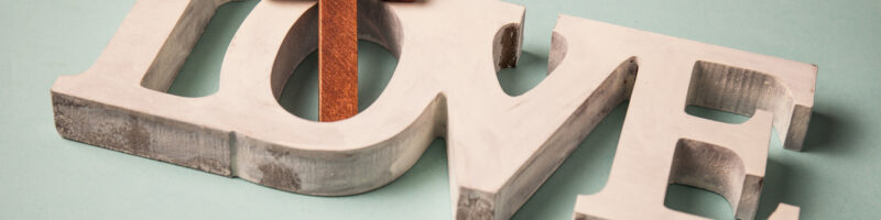 White wooden letters spell LOVE with a rusty brown cross as the O on a pale turquoise background.