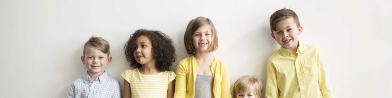 Five children stand in a line against a light wall, wearing yellow and pastel clothing and smiling.